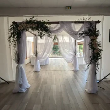 Houppa de mariage en bois décorée de voilages blancs et de compositions florales suspendues, installée en intérieur pour une cérémonie élégante.