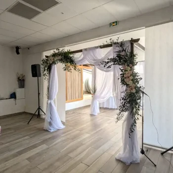 Vue latérale d’une houppa de mariage en bois avec drapés blancs et guirlandes florales pastel, en location par Iliade Location, installée dans une salle de réception.