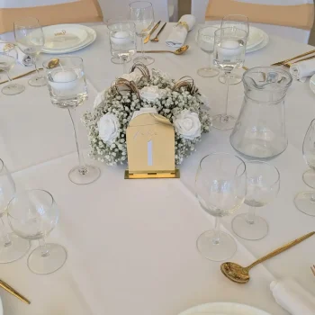 Table de mariage élégante décorée en blanc et or, centre de table fleuri avec gypsophile et roses blanches, numéro de table doré, vaisselle raffinée et couverts dorés.