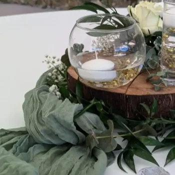 Photophore boule ou sphère pour bougie - Décoration de mariage dans le Var : Une décoration de table élégante et naturelle avec un photophore en forme de boule en verre, contenant une bougie flottante. Le photophore est posé sur une base en bois, entouré de feuillage vert, de gypsophiles et d'une serviette en tissu vert. Ce décor rustique et raffiné évoque un mariage en plein air, avec une ambiance champêtre et romantique.