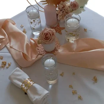 Trio de photophores en verre avec bougies flottantes - Décoration de mariage : Une table de mariage élégante décorée de trois photophores en verre, chacun contenant une bougie flottante. Autour des photophores, un chemin de table en satin beige se déroule, accompagné d'une serviette roulée avec un anneau doré. Des éléments décoratifs comme des étoiles scintillantes, des roses en soie, et des touches d'or sont répartis sur la nappe blanche, créant une ambiance douce et romantique.